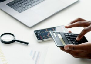 pessoa usando calculadora, mesa com notebook, folha, lupa e celular, ilustrando uma analise de concorrencia