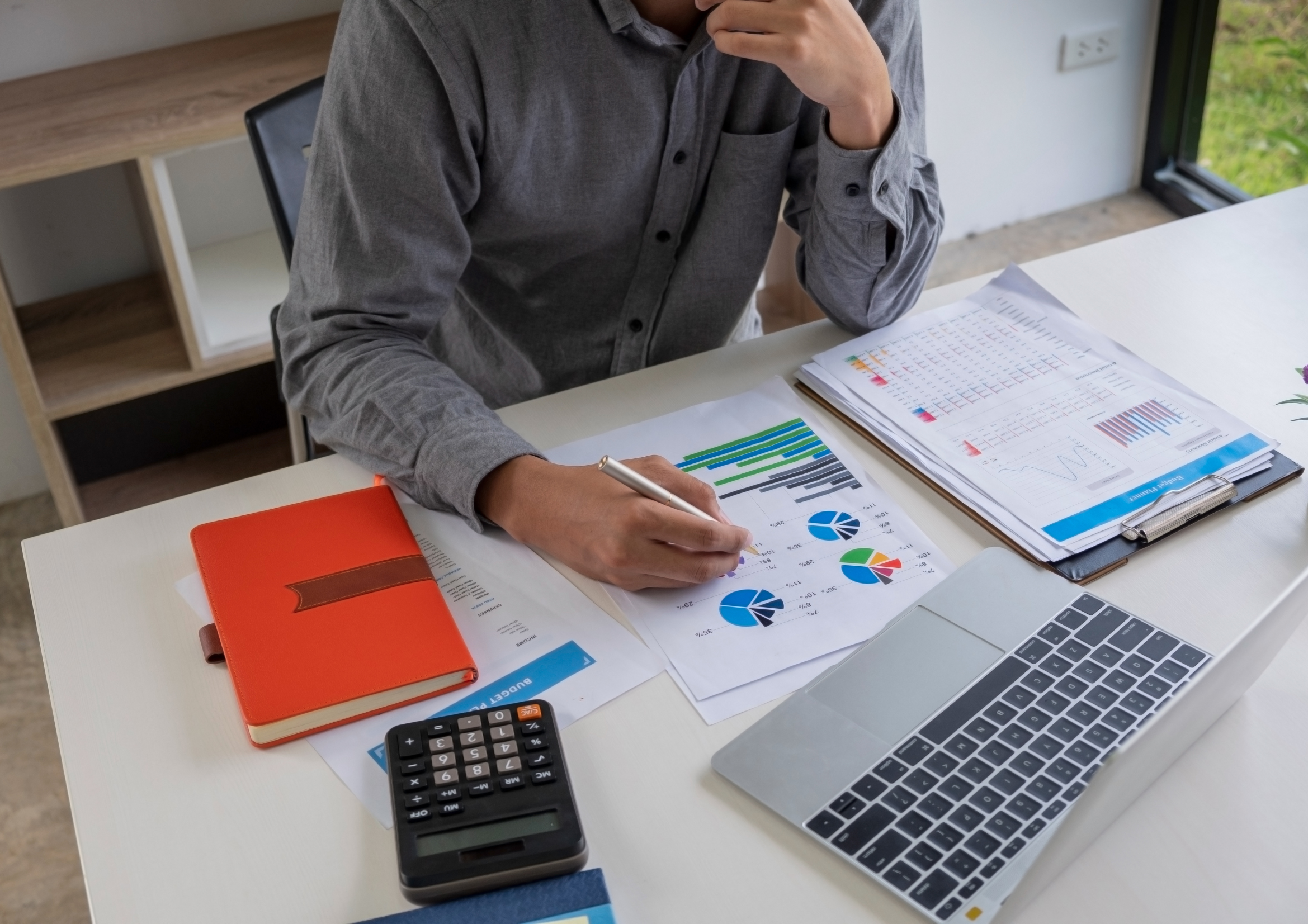 Pessoa fazendo análise, na mesa com gráficos, caderno, computador e calculadora, para ilustrar o processo de pricing estratégico